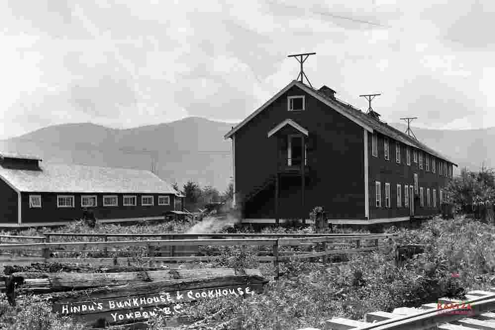 A black-and-white photograph displaying the large bunkhouse and cookhouse buildings at the Industrial Timber Mills Ltd, Youbou, B.C. There is a sign that reads: “Hindu’s Bunkhouse & Cookhouse” and part of a railway in the foreground.